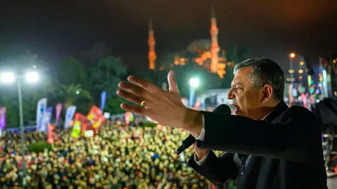 Özgür Özel bir meydanda otobüsün üstünden konuşurken elini kaldırmış, arkaplanda çok sayıda kişinin meydanı doldurarak kendisini dinlediği görülüyor