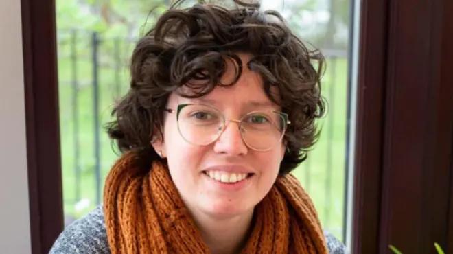 Jenny Watt smiles while looking directly into the camera. She has glasses, curly dark hair and an orange/brown scarf wrapped around her neck.
