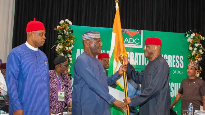 David Mark wey wear black cloth and red cap hold di flag with Atiku Abubakar wey wear blue cloth wit cap wey get designs. ADC Party banner and oda pipo dey for di background