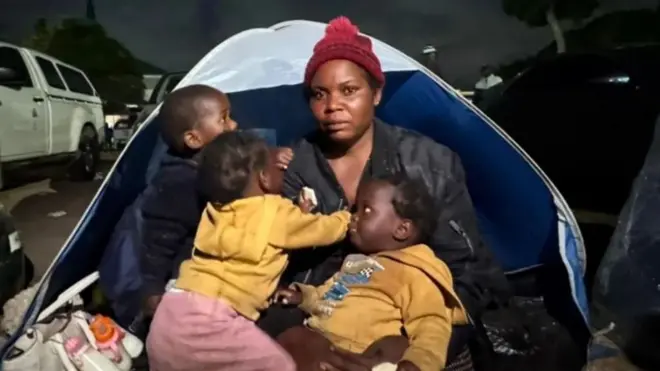 Esnat Joseph, yambaye inkofero y’ubwoya y’ibara ritukura, yambaye kandi ikoti yirabura, yicaye imbere y'ihema ari kumwe n'abana biwe batatu b'amahasa