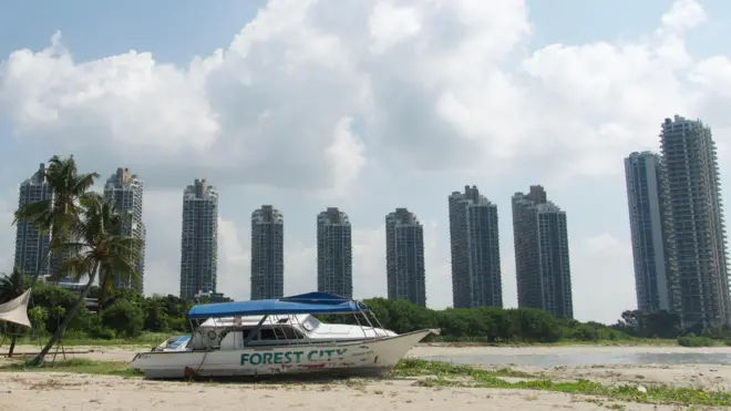 Un barco destartalado con la palabra "Forest City" en su costado descansa sobre la arena de una playa, junto a un montón de escombros y palmeras, con edificios altos al fondo.