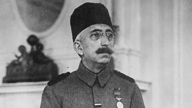 Retrato del sultán Mehmed VI. Tiene puestos lentes y un tarboosh en la cabeza. Viste un uniforme y le cuelga una medalla en el lado izquierdo de su pecho 