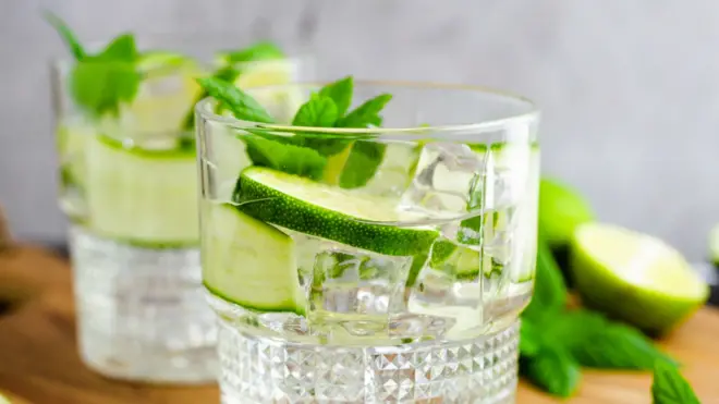 Two glasses of gin and tonic with sliced lime and mint, resting on a wooden board. Sliced lime and mint leaves are lying next to the glasses.