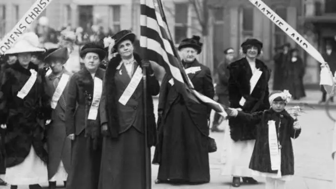 Una foto en blanco y negro de 1909 en el que se ve a mujeres marchando en favor de la prohibición del alcohol en EE.UU. Una en la mitad lleva una bandera estadounidense, la cual es desplegada por una niña- 