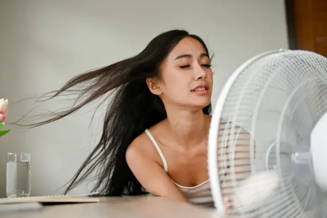 A beautiful Asian woman, feeling overheated, cools herself down with an electric fan
