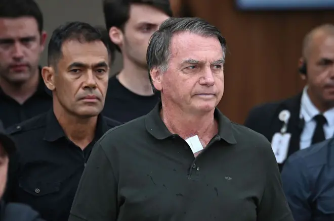 Jair Bolsonaro usando uma camisa preta e com um curativo no pescoço ao lado de policiais