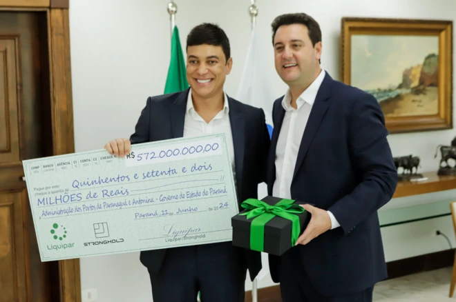 Foto com Cleiton Santos Santana à esquerda, o governador Ratinho Jr. à direita, ambos usando ternos escuros e sem gravatas com Cleiton segurando um cheque simbólico de R$ 572 milhões