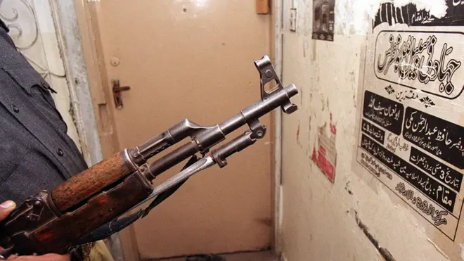 عکسی از یک سلاح کلاشنیکوف و دیواری که در آن مضامین جهادی نوشته شده استږ