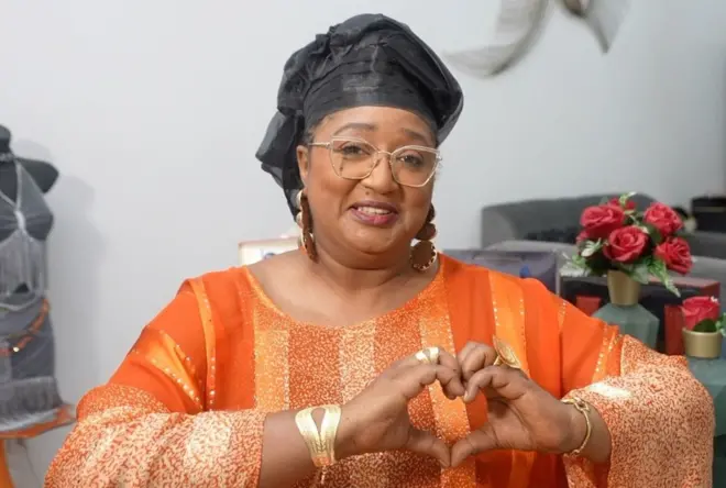 Une femme portant un boubou orange avec des rayures verticales et un foulard noir sur la tête. Elle croise ses doigts en forme de coeur et sourit à la caméra.