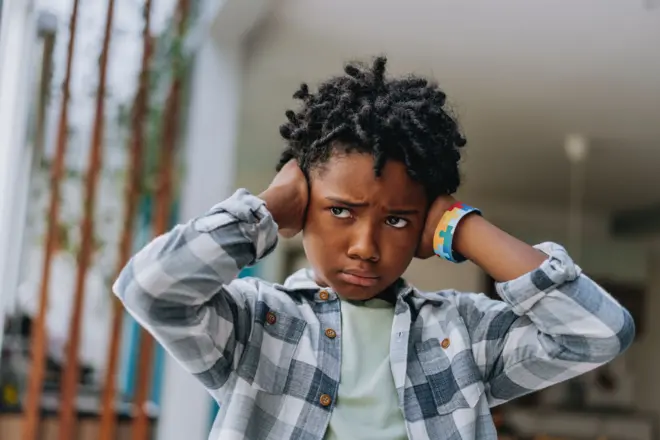 Un enfant porte une chemise carrelée blanc et noir avcec un t-shirt vert en dessous, se couvre les deux oreilles avec ses mains, il a des cheveux afro et un visage en colère.