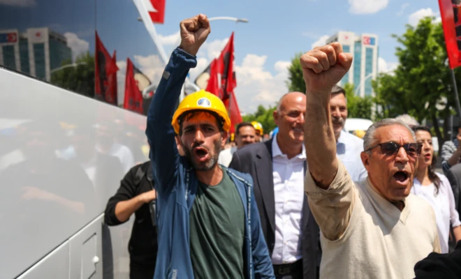 Sarı kasklı bir madenci ve yanındakiler sağ kolllarını havaya kaldırmış ve yumruk yapmış. Slogan atarak yürüyorlar.
