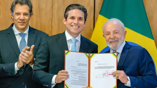 Haddad, Motta e Lula sorrindo e posando para foto, lado a lado; Motta e Lula seguram documento do projeto de isenção