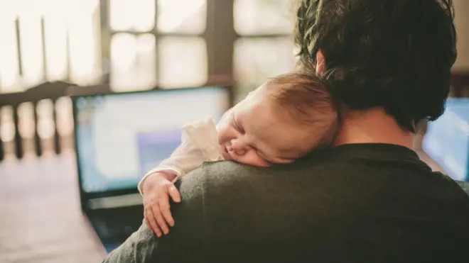 Father working on his laptop while his newborn baby girl sleeps on his shoulder