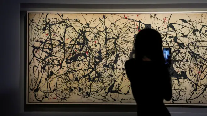 A person with long hair in silhouette is seen facing the artwork holding their mobile phone to video the painting. The artwork is long horizontally and the grey canvass appears a light tan colour under the gallery lights. It features black paint flicks and splashes all over it, together with some fewer red markings.