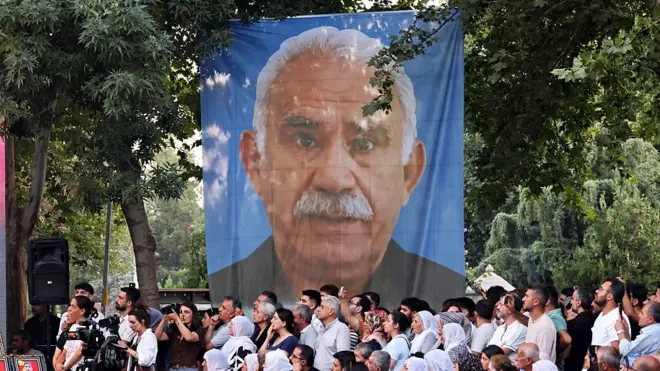 Öcalan'ın mavi bir zeminde beyaz saçlı büyük bir poster gibi fotoğrafı asılmış. Ağaçlık alanda yapılan bu etkinlikte çok sayıda kişi bir yere doğru bakıyor