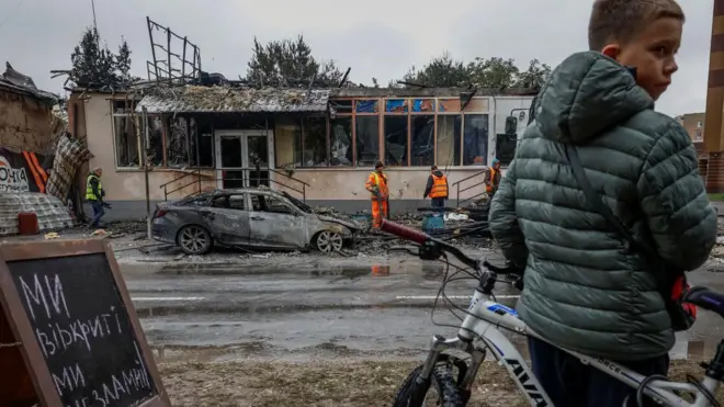 Мальчик в зелёной куртке с велосипедом на фоне разрушенного здания и сгоревшего автомобиля. В кадре видны спасатели в жёлтых и оранжевых жилетах