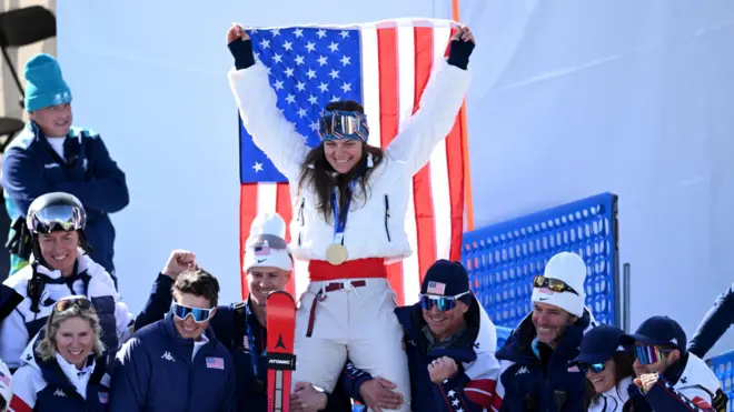 Бризи Джонсон с золотой медалью держит флаг США в окружении команды.