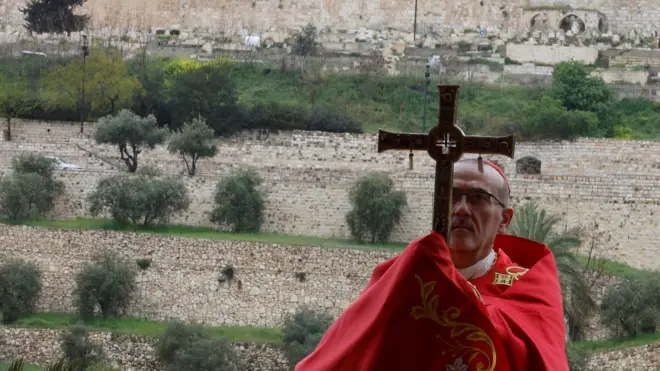 Пиццабалла в красном облачении с крестом в руках на фоне стен.