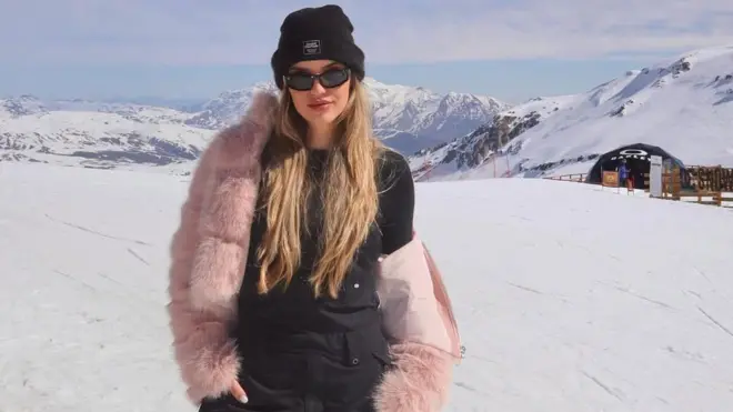 Джуліана Пассареллі стоїть у засніжених горах у рожевій хутряній куртці та темному одязі. У неї довге світле волосся, вона дивиться прямо в камеру, тримаючи руки в кишенях