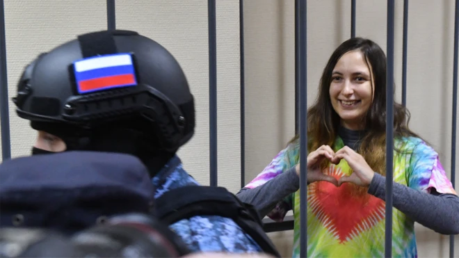Саша Скочиленко показывает руками сердце, находясь за решеткой. 