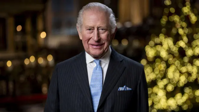 Король Чарльз в темному смугастому костюмі з блакитною краваткою та хустинкою у нагрудній кишені стоїть у великому, урочистому приміщенні. У тлі – м’яко розмиті теплі вогники та різдвяна ялинка, що створюють святкову атмосферу.
