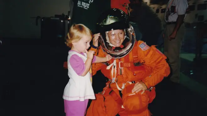 A astronauta Eileen Collins veste um traje espacial laranja e capacete transparente. Ela sorri ao se agachar ao lado da filha, de três anos. A menina veste roupa rosa e branca, leva um dedo à boca e apoia a outra mão no capacete da mãe.