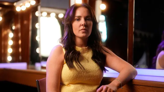 Uma mulher vestindo uma blusa amarela texturizada sem mangas, sentada em frente a um espelho com luzes redondas de maquiagem em um camarim.