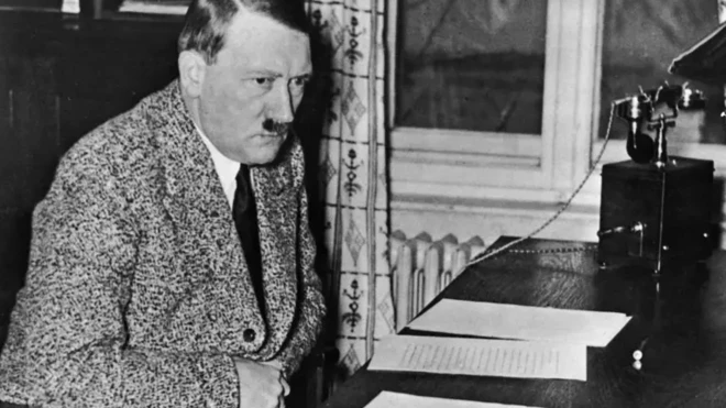 هیتلر با لباس غیرنظامی پشت میزی نشسته و چند دست نوشت جلوی اوست به چیزی خیره شده است