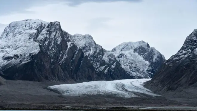 A imagem mostra montanhas com neve por cima, sob um céu cinza. 