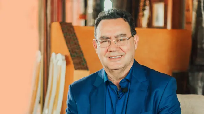 Um homem de meia-idade, Augusto Cury está sentado em um ambiente interno com iluminação quente, vestindo um terno azul sobre uma camisa azul mais escura e óculos de armação fina, sorrindo; ao fundo, vê-se uma luminária acesa e móveis de madeira.