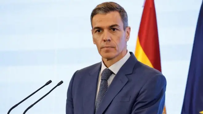 Nötr bir ifadeye sahip, kısa ve griye dönmüş kahverengi saçlı Pedro Sanchez, bir basın toplantısı sırasında dinlerken takım elbise ve kravatlı.