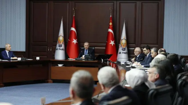 Cumhurbaşkanı Erdoğan masa başında, çevresinde parti yetkilileri. Türkiye ve AKP bayrakları