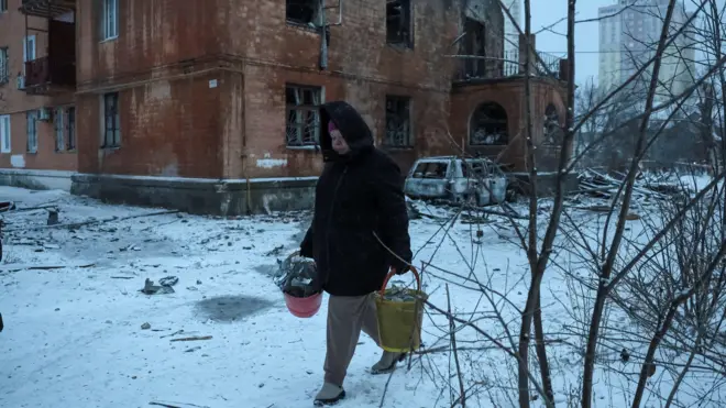 Жінка несе відра з уламками на тлі пошкодженого обстрілом будинку, на землі лежить сніг