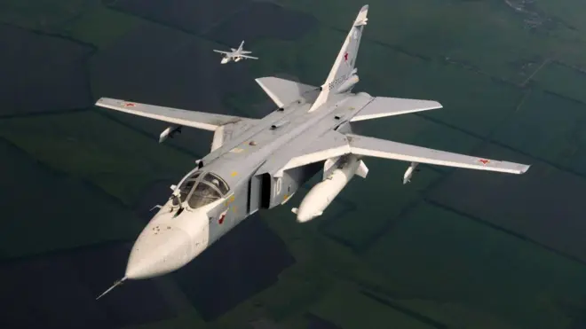 Військовий літак сіро-білого кольору, зафіксований у польоті над зеленими полями. Літак має розкладні крила змінної стрілоподібності, підвішені паливні баки або боєприпаси та червоні зірки на крилах і фюзеляжі. Позаду, на відстані, летить ще один подібний літак.