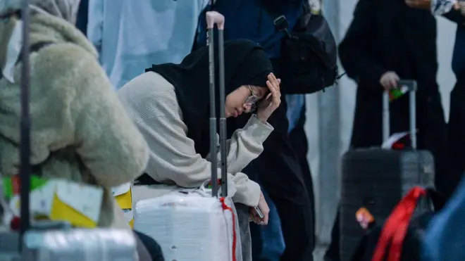 Seorang perempuan muda berhijab sedang duduk dengan wajah lesu dan mata terpejam dengan tangan kirinya memegang kepala di tengah banyak orang.