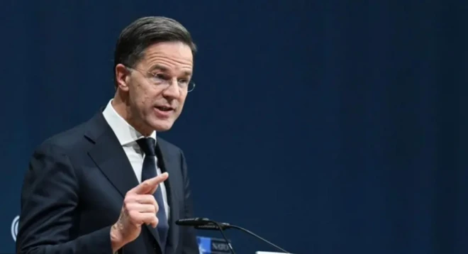 Takım elbiseli ve gözlüklü bir erkek elini kaldırmış mavi bir fonda konuşuyor