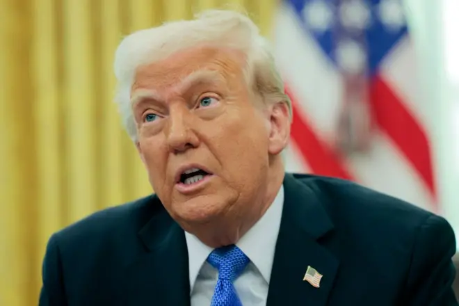 ترامب يرتدي بدلة رسمية كحلية وربطة عنق زرقاء وعلى جاكيت البدلة دبوس يحمل العلم الأمريكي. وخلفه العلم الأمريكي.