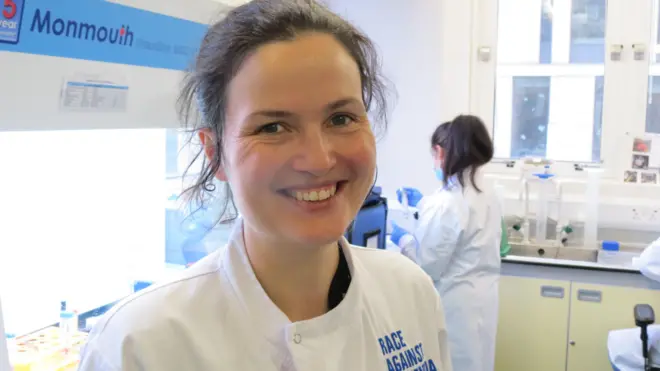 A smiling scientist with tied back hair looks to camera in her laboratory. She is wearing a white lab coat with the words "Race Against" clearly written and the word dementia can be inferred. There are people at work behind her.