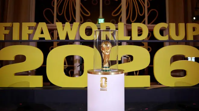 Di World Cup on display on white stand prior to di draw in December in Washington wit Fifa World Cup 2026 in big gold letters for di background.