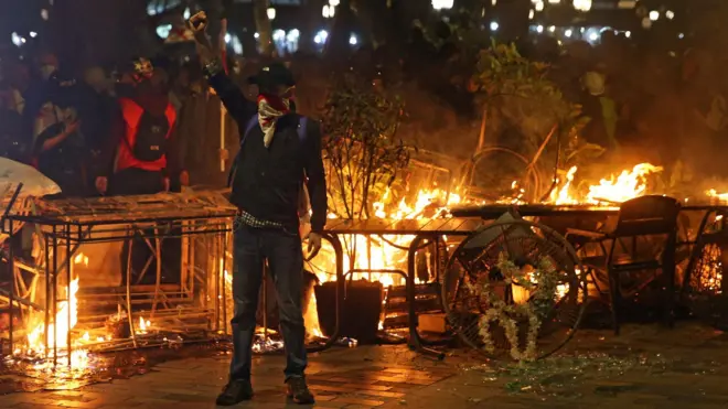 Демонстрант стоит с поднятым кулаком на фоне горящей баррикады