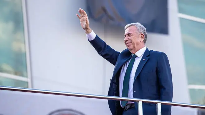 Mansur Yavaş bir seçim otobüsü üzerinde mavi ceketiyle sağ elini kaldırarak halkı selamlıyor. 