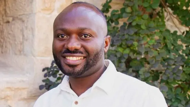 Ifoto ngufi igaragaza Dr Célestin Mutuyimana yambaye ishati y'umweru arimo kumwenyura. Inyuma ye hari ibyatsi n'urukuta rw'inzu y'amabuye. 