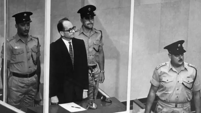 آدلف آیشمن در حصار شیشه‌ای در دادگاه ایستاده است