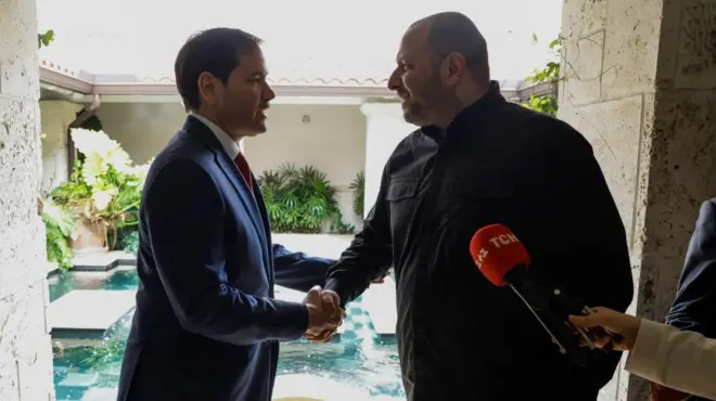 大柄で薄い口ひげをたくわえ黒いシャツ姿のウメロフ氏と、スーツ姿のルビオ氏がお互いに向き合い、笑顔で握手している。明るい中庭や池を背にしているため、逆光になっている。手前には、赤いマイクとそれを差し出す記者の右手が写っている