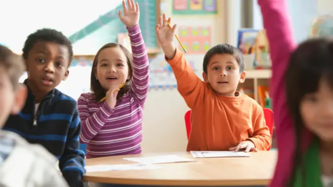 A boy and a girl sit next to each other and both raise one hand in the air. The girl wears a purple and white stripy top, while the boy is in orange. They are surrounded by peers of a similar age in the foreground.