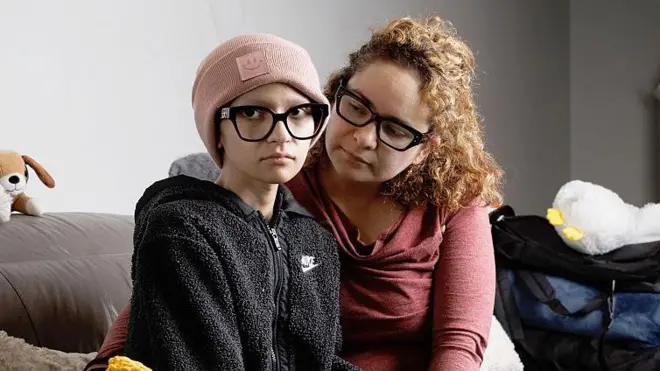 Ofelia, delgada y luciendo un gorro, mira a la cámara con una expresión de tristeza mientras su madre la mira y la rodea con sus brazos.