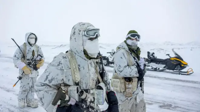 Uma imagem mostrando três militares canadenses em uma paisagem nevada, vestindo uniformes de camuflagem branca, máscaras faciais, luvas e segurando vários tipos de munição enquanto caminham para a direita da imagem. Atrás deles, vê-se uma moto de neve estacionada.