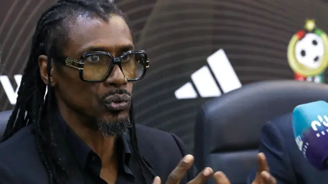 Aliou Cisse porte de grosses lunettes à monture noire alors qu'il parle dans un microphone lors d'une conférence de presse avec le logo de la Fédération libyenne de football en arrière-plan.
