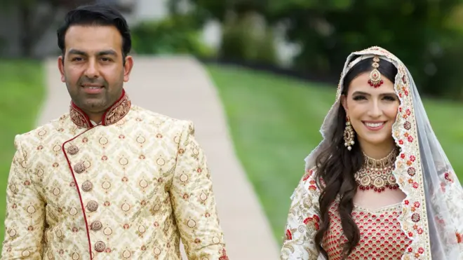 Sammi e Safi posam de mãos dadas em traje de casamento. Ela usa um vestido afegão tradicional de noiva, branco e vermelho, com lenço na cabeça; ele veste um sherwani, traje dourado típico de noivos.