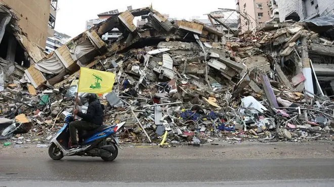 یک موتورسوار با پرچم حزب‌الله لبنان از کنار ویرانه‌های ساختمان‌ها می‌گذرد